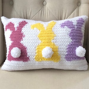 May include: A white crocheted pillow with three bunny silhouettes in pink, yellow, and purple. Each bunny has a white pom-pom tail.