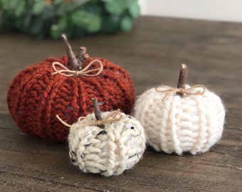 May include: Three knitted pumpkins in various sizes. The largest pumpkin is a deep orange color, the medium pumpkin is white, and the smallest pumpkin is a cream color. All three pumpkins have a rustic twig stem and a twine bow.