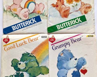 May include: Four Butterick sewing pattern envelopes featuring Care Bears characters: Cheer Bear in pink with a rainbow, Friend Bear in orange with flowers, Good Luck Bear in green with a clover, and Grumpy Bear in blue with a rain cloud and heart.