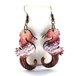 May include: A pair of wooden seahorse earrings with a pink and white watercolor design. The earrings have bronze hooks.