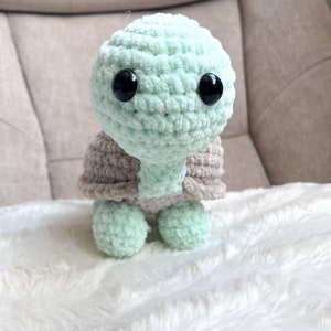 K&ouml;nnte beinhalten: Ein geh&auml;keltes gr&uuml;nes Pl&uuml;sch-Schildkr&ouml;tenspielzeug mit schwarzen Augen. Die Schildkr&ouml;te tr&auml;gt eine braune geh&auml;kelte Schale.