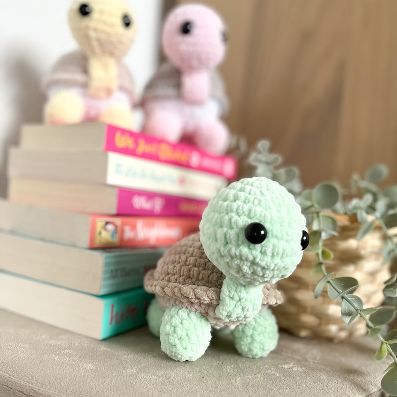 K&ouml;nnte beinhalten: Ein geh&auml;keltes gr&uuml;nes Schildkr&ouml;tenspielzeug mit brauner Schale, das auf einem Stapel B&uuml;cher liegt. Die Schildkr&ouml;te hat schwarze Augen und eine weiche, flauschige Textur.