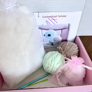 Puede incluir: Un kit de amigurumi de tortuga de peluche de crochet en una caja rosa. El kit incluye dos ovillos de hilo, uno verde claro y uno marr&oacute;n, una bolsa de relleno, dos agujas de crochet y una bolsa de organza rosa. La caja tambi&eacute;n incluye una hoja de instrucciones impresa con una foto de la tortuga terminada.