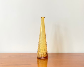 May include: A tall, slender, amber-colored glass vase with a conical shape and a textured, grid-like pattern of raised squares covering its exterior surface. The vase features a narrow neck and a small, circular opening at the very top.