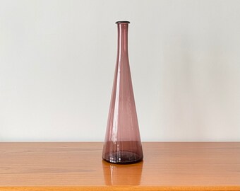 May include: A tall, slender, conical glass vase in a translucent mauve purple hue. The vase features a wide, flat circular base that tapers upwards into a long, narrow neck with a simple, smooth rim at the top, showcasing a clean, minimalist form.