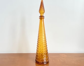 May include: A tall, amber-colored glass decanter with a conical shape and a matching pointed stopper. The surface features a repeating hobnail or dot pattern that covers the entire body of the bottle and the stopper, creating a textured finish.
