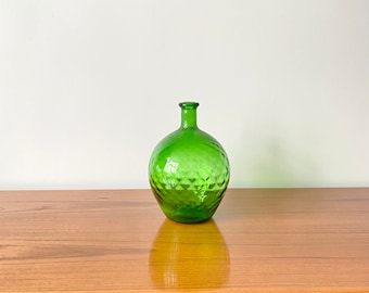 May include: A round, green glass bottle with a narrow neck and a textured, diamond-patterned surface. The glass is translucent, allowing light to pass through the vessel, which features a small, circular opening at the top and a rounded base.