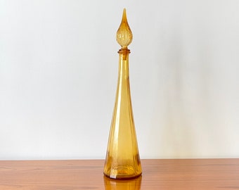 May include: A tall, amber-colored glass decanter with a conical body and a slender neck. The decanter features a matching amber glass stopper with a teardrop-shaped finial that has vertical ribbing, creating a sleek and elongated silhouette.
