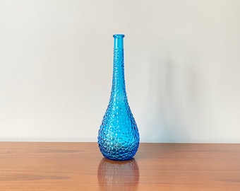 Peut inclure: Un vase en verre bleu, haut et élancé, doté d'un col étroit et d'une base bulbeuse. La surface extérieure du vase est recouverte d'un motif texturé composé de bulles circulaires en relief de tailles variées, offrant un aspect translucide.