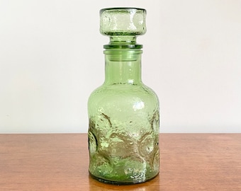 May include: A light green glass decanter featuring a textured, dimpled surface and a matching cylindrical glass stopper. The vessel has a rounded body that tapers into a narrow neck, with a thick, clear glass rim at the top of the stopper.
