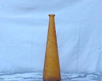 May include: A tall, amber-coloured glass bottle with a conical shape. The exterior surface features a textured, vertical ribbed pattern that runs from the base up to the narrow neck, creating a distinct tactile appearance in the light.