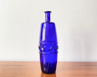 May include: A tall, cylindrical cobalt blue glass bottle featuring a narrow neck and a distinct decorative wavy glass band encircling the center of the body. The bottle has a smooth, translucent finish and a flat base, standing upright with a uniform color.