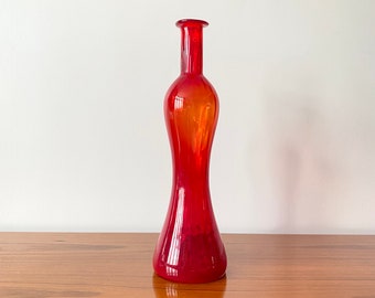 May include: A tall, slender, translucent red glass vase with a narrow neck and a flared, wide base. The glass features a smooth, polished surface that catches the light, creating subtle variations in the vibrant red colour throughout the body.