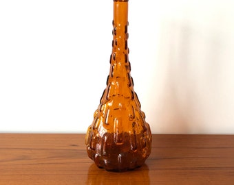 May include: A tall, amber-colored glass bottle with a narrow neck and a bulbous base. The exterior surface features a repeating, raised rectangular grid pattern that creates a textured, tactile appearance across the entire body.