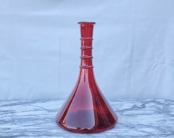 May include: A deep red glass decanter featuring a wide, conical base that tapers into a slender, elongated neck. The neck is accented with three distinct horizontal ringed ridges, and the opening at the top is finished with a flat, circular rim.