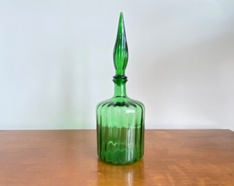 May include: A translucent emerald green glass decanter featuring a cylindrical body with vertical ribbed detailing and a matching tall, pointed, teardrop-shaped glass stopper that fits securely into the narrow neck of the vessel.