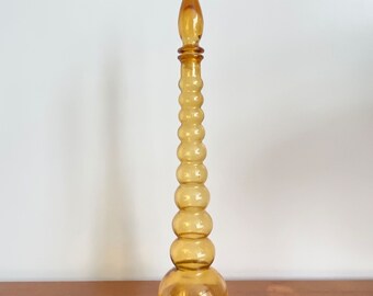 May include: A tall, amber-colored glass decanter featuring a rounded base and a long, slender neck composed of a series of stacked, graduated spherical segments, topped with a pointed, teardrop-shaped glass stopper.