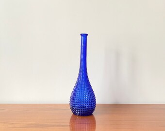 May include: A tall, slender cobalt blue glass vase featuring a textured hobnail pattern across its bulbous base and elongated neck. The vase has a narrow opening at the top and a rounded bottom, showcasing a vibrant, translucent blue color throughout.
