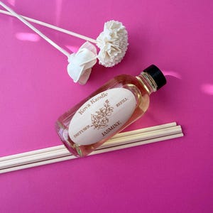 May include: A clear glass bottle of Kova Kandle jasmine diffuser refill oil with a black cap and an oval label. The bottle is accompanied by several straight reed sticks and two white decorative flower-shaped reed diffusers on a pink surface.