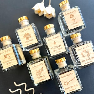 May include: An assortment of Kova Kandle reed diffusers in clear glass bottles with wooden labels and gold-colored caps. The labels feature various scents like "Red Rose," "Pine Tree," and "Pink Cashmere." White flower-shaped diffusers are also visible.