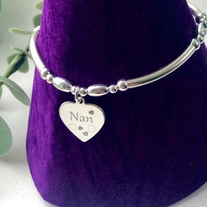 May include: A silver bracelet featuring a heart-shaped pendant engraved with the word "Nan" and heart motifs. The bracelet has a tubular design with small, round beads. The bracelet is displayed on a purple velvet surface.