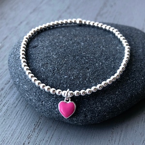 May include: A silver bracelet with a pink heart charm. The bracelet is made of small, round silver beads and features a bright pink heart pendant. The bracelet is displayed on a dark grey stone surface.