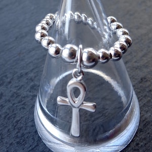 May include: A silver beaded ring with a dangling ankh charm. The ring is made of small, round beads and the ankh charm is a classic Egyptian symbol. The ring is displayed on a clear, cone-shaped stand against a dark background.
