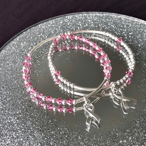 May include: Two silver bracelets with pink crystal beads and silver ribbon charms. The bracelets are displayed on a reflective, glittery surface, with a dark purple background. The bracelets are a delicate and elegant accessory.