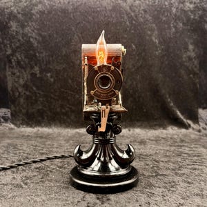 May include: A unique steampunk-style lamp crafted from a vintage camera. The lamp features a black base with an ornate design, a central camera body, and a decorative light bulb. The lamp is connected to a black cord.