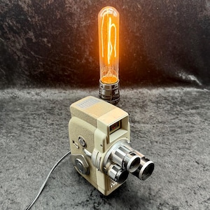 May include: A vintage-style movie camera lamp featuring a beige body and a dual-lens turret. A tall, cylindrical Edison-style light bulb with a glowing orange filament is mounted on top of the camera, connected by a black power cord.