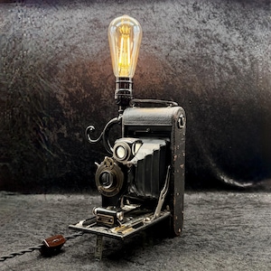 Peut inclure: Un appareil photo pliant noir vintage reconverti en lampe de table. L'objet présente un corps texturé façon cuir, un mécanisme à soufflet et un objectif métallique. Une ampoule de style Edison est fixée au sommet, diffusant une lueur chaude.
