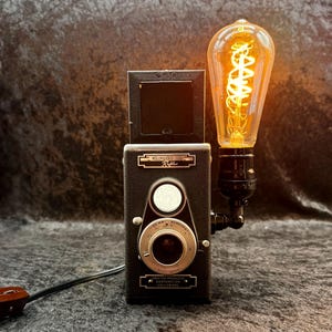 Może przedstawiać: Zabytkowy czarny aparat Hollywood Reflex przerobiony na lampę, wyposażony w świecącą żarówkę w stylu Edisona z widocznym spiralnym żarnikiem przymocowanym z boku. Na korpusie aparatu widnieje napis Hollywood Reflex oraz Crafter Products Corporation.
