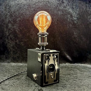 May include: A table lamp constructed from a vintage black box camera. The lamp features a round, amber-tinted Edison-style light bulb with visible filaments mounted on top. The camera body has a silver-toned faceplate with geometric detailing.