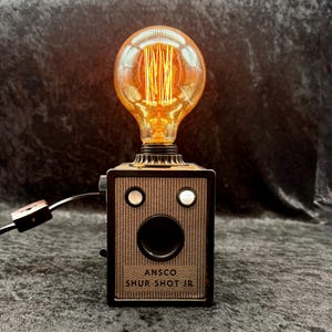 Puede incluir: Una lámpara de estilo vintage con una bombilla Edison brillante encima de una cámara ANSCO Shur Shot Jr. reutilizada. La lámpara tiene una combinación de colores marrón y negro, con el texto "ANSCO SHUR SHOT JR." en el cuerpo de la cámara.