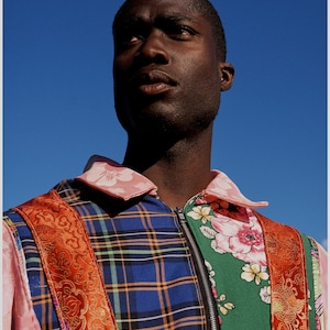 May include: A man wearing a multi-colored patchwork jacket with a zipper closure. The jacket features a plaid pattern, floral print, and a red and gold patterned fabric.