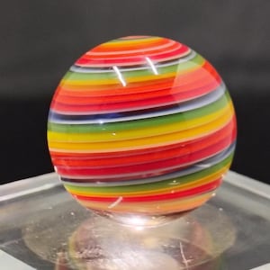 May include: A colorful glass marble with a spiral design. The marble features bands of red, orange, yellow, green, blue, and white. The glass is smooth and reflective, with a glossy finish. The marble is sitting on a clear surface.