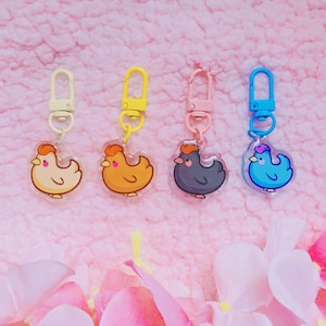 May include: Four acrylic keychains featuring cartoon chicken designs in cream, orange, dark grey, and blue, each attached to a matching coloured plastic lobster clasp. The chickens have simple rounded bodies, small beaks, and pink cheek accents.