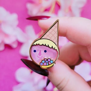 May include: A gold-colored enamel pin shaped like an ice cream cone with a face. The pin features a pink face, a waffle cone hat, and a yellow hair-like trim. The character is holding a small bowl of colorful round candies.
