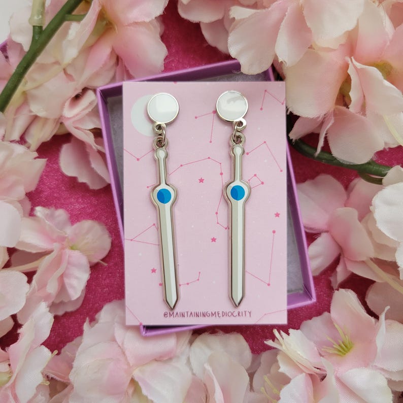 May include: Silver-colored sword-shaped earrings with a blue center detail and a white top. The earrings are displayed on a pink card inside a purple box, surrounded by pink flowers. The text "@MAINTAININGMEDIOCRITY" is at the bottom of the card.