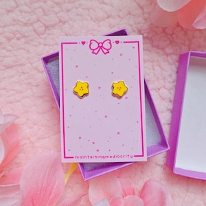 May include: A pair of yellow star-shaped stud earrings displayed on a light pink rectangular card with a bow graphic and the text maintainingmediocrity. The earrings are presented inside an open purple box with a light gray interior lining.