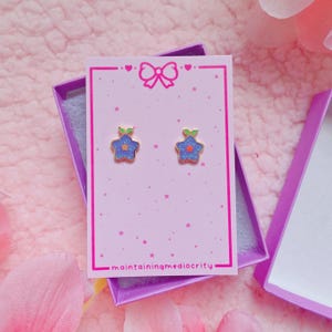 May include: A pair of star-shaped stud earrings with blue glitter enamel, a small orange center, and two green leaves at the top. The earrings are displayed on a light pink card featuring a bow icon and the text maintainingmediocrity.