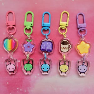 May include: A collection of five acrylic keychains with colorful designs. Each keychain features a different charm: a rainbow heart, a purple flower, purple shorts, a coffee mug, and a yellow star. Each keychain has a matching color clasp and a small apple charm.