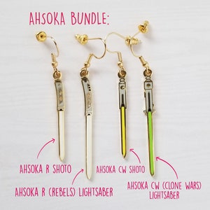 Puede incluir: Un conjunto de tres pendientes dorados con los sables de luz de Ahsoka Tano de la franquicia Star Wars. Los pendientes tienen la forma de los sables de luz, con hojas blancas, amarillas y verdes. El texto "AHSOKA BUNDLE" es visible en la parte superior de la imagen.
