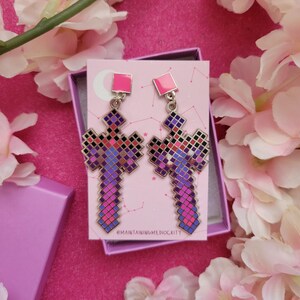 May include: A pair of pixelated cross earrings with pink and purple hues. The earrings have a square top and a cross-shaped design with a pixelated pattern. The earrings are displayed on a pink card inside a pink box, surrounded by pink flowers.