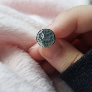May include: A silver-toned enamel pin featuring a black and white illustration of the Death Star from Star Wars.