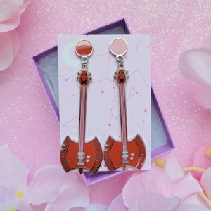 May include: A pair of guitar-shaped earrings with a red-orange body and silver accents. The earrings are displayed on a pink card inside a purple box, surrounded by pink flowers. The earrings have a cartoonish design.