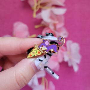 May include: A purple cartoon mouse holding a slice of pizza with pepperoni, green peppers, and mushrooms. The pizza slice is yellow with a red crust. The mouse is holding the pizza with its front paws and has a curled tail. The pin is gold-toned.