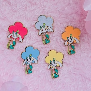 May include: Five enamel pins featuring a cartoon character with a flower head and a heart-shaped leaf. The pins are in different colors: light blue, yellow, orange, pink, and light blue with glitter.