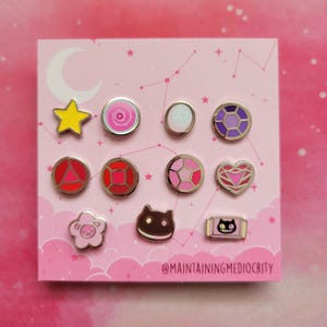 May include: A collection of enamel pins with various designs, including a yellow star, a pink rose, and a purple gem. The pins are arranged on a pink card with a constellation design and the text "@MAINTAININGMEDIOCRITY".