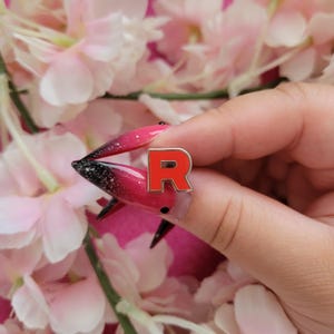 May include: A red enamel pin in the shape of the letter "R" with a silver border. The pin is held against a background of pink flowers. The pin is a small accessory.